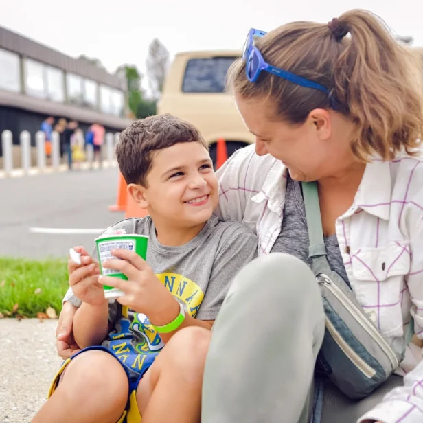 A young boy sits with a woman in a purple and white plaid shirt. The boy is eating ice cream out of a cup with a spoon. The two are looking at each other and smiling.