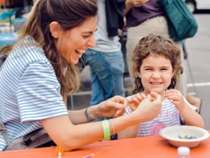 A woman laughing with a toddler girl as they make bracelets together.
