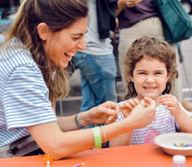 A woman laughing with a toddler girl as they make bracelets together.
