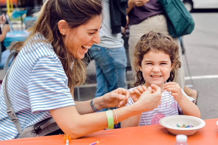 A woman laughing with a toddler girl as they make bracelets together.