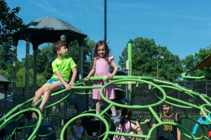 A group of children playing on a playground. A boy sits on top of the bars and a girl stands on top of it.