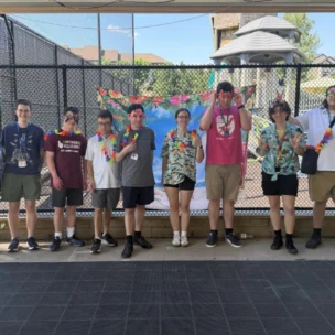 A group of nine people stand in a row outside by a playground, wearing leis and summer clothes, posing and smiling for the camera.