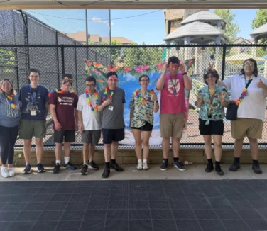 A group of nine people stand in a row outside by a playground, wearing leis and summer clothes, posing and smiling for the camera.