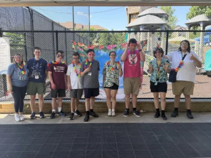 A group of nine people stand in a row outside by a playground, wearing leis and summer clothes, posing and smiling for the camera.