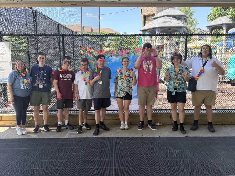 A group of nine people stand in a row outside by a playground, wearing leis and summer clothes, posing and smiling for the camera.