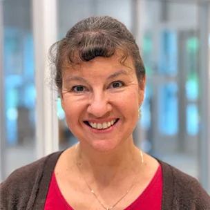 Woman smiling at the camera, wearing a red top and brown cardigan, standing indoors with blurred glass doors in the background.