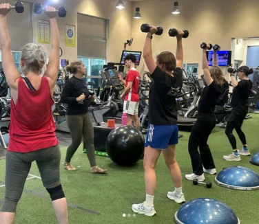 Women in a gym lifting dumbbells over their heads.