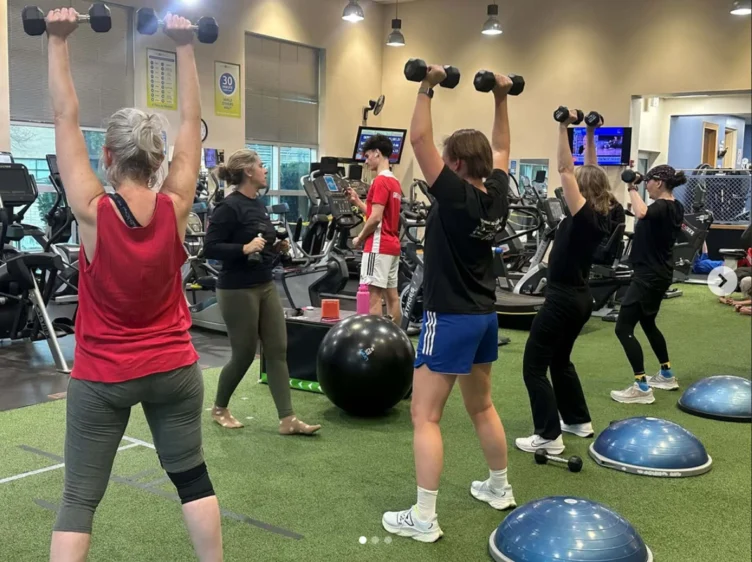 Women in a gym lifting dumbbells over their heads.