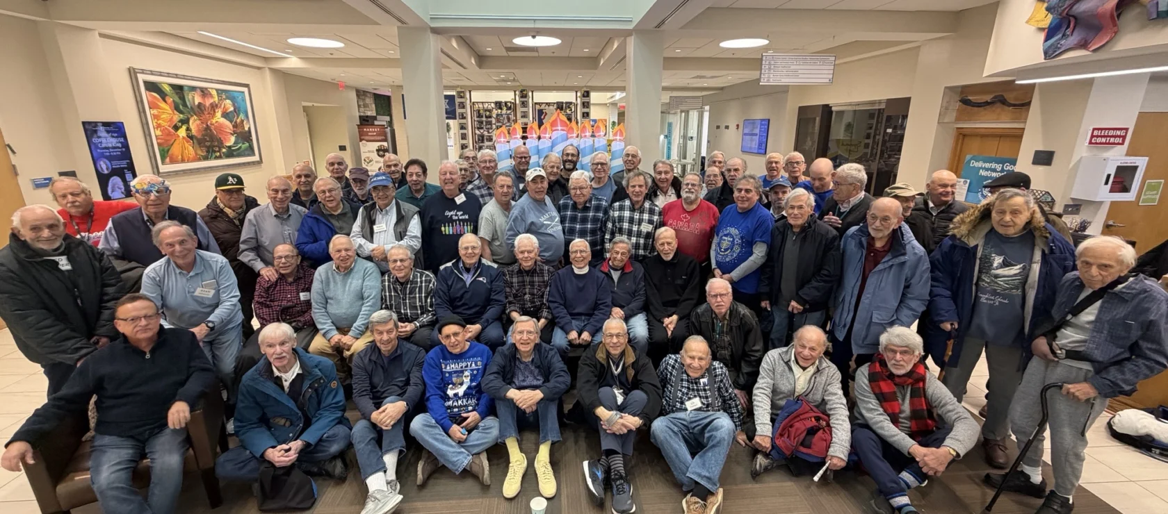 A large group of older men pose together for a photo in a well-lit lobby with artwork and signs on the walls.