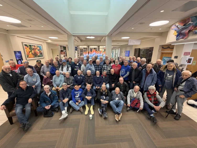 A large group of older men pose together for a photo in a well-lit lobby with artwork and signs on the walls.