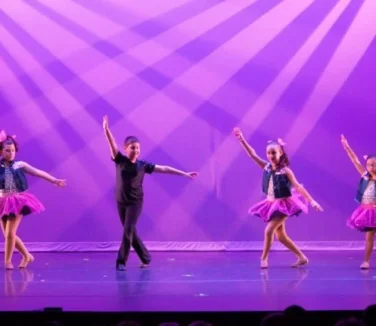 Five young girls and a boy in the middle posing onstage in costumes.
