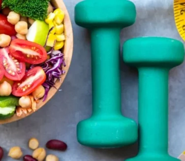 A healthy bowl of salad with dumbbells and a measuring tape.