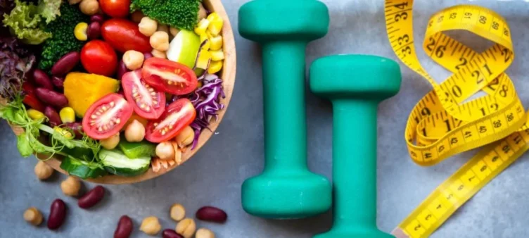 A healthy bowl of salad with dumbbells and a measuring tape.