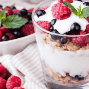 A glass cup filled with raspberries, blueberries, granola, and yogurt. A bowl of fruit sits in the background while a spoon resides beside the cup.