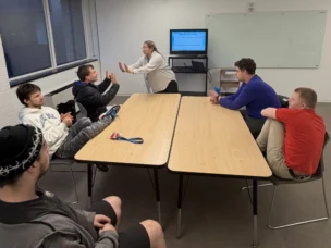 Six people sit around two tables in a meeting room while one person stands in front of a monitor, speaking and gesturing with their hands.
