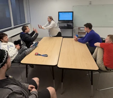 Six people sit around two tables in a meeting room while one person stands in front of a monitor, speaking and gesturing with their hands.