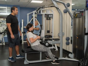 A trainer oversees a young woman using gym equipment.
