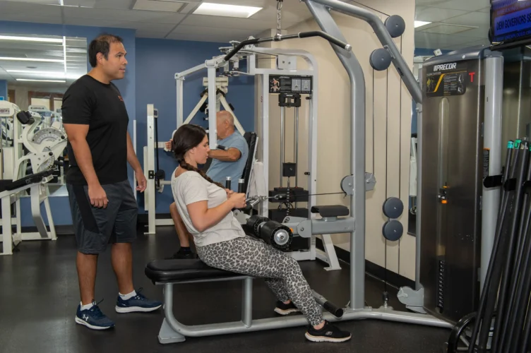 A trainer oversees a young woman using gym equipment.