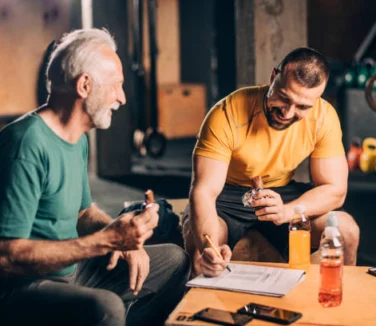 An older man and a younger man sit in a gym, eating protein bars and smiling while reviewing notes or a workout plan on a clipboard.