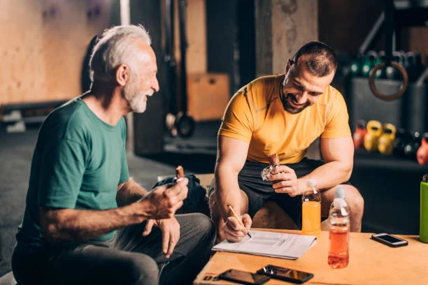 An older man and a younger man sit in a gym, eating protein bars and smiling while reviewing notes or a workout plan on a clipboard.