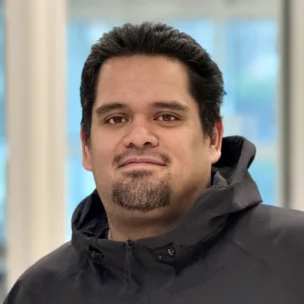 A man with short dark hair, a goatee, and a black jacket stands indoors in front of a blurred window background.
