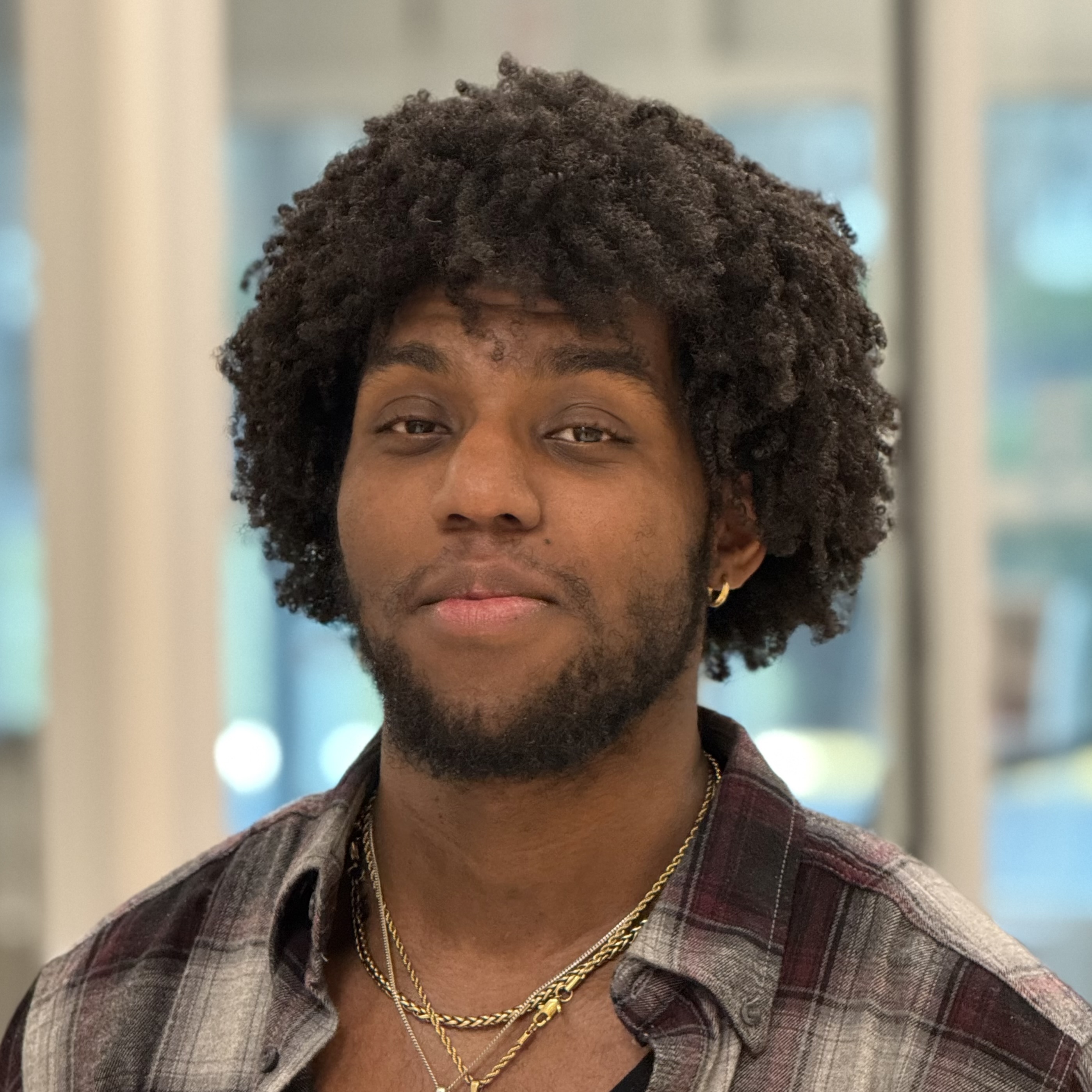 A young man with curly hair, a short beard, wearing a plaid shirt, gold chains, and a small gold hoop earring, stands indoors in front of large windows.