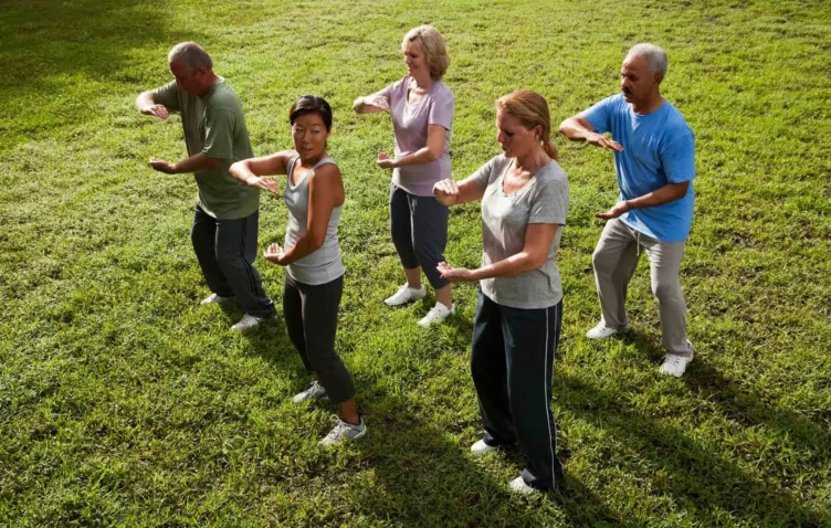 Five adults practice tai chi together outdoors on a grassy field, standing in a row and performing synchronized movements.