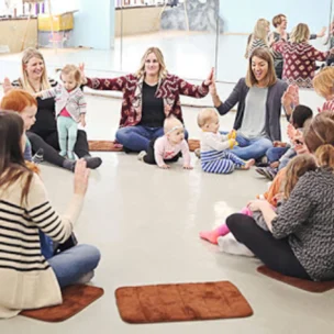 A group of parents and young children sit in a circle on mats in a bright room, participating in an interactive group activity.