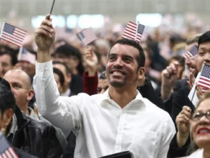 A large group of people, many holding small American flags, attend an indoor event; most appear to be looking forward and smiling.