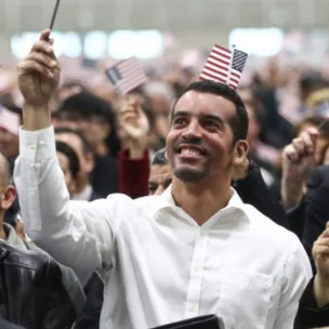 A large group of people, many holding small American flags, attend an indoor event; most appear to be looking forward and smiling.