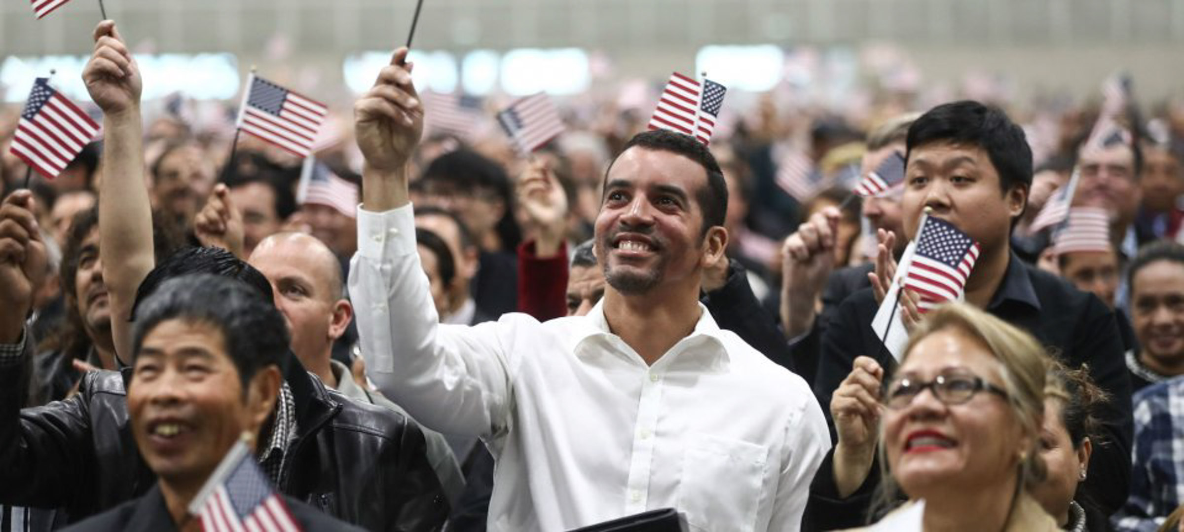 A large group of people, many holding small American flags, attend an indoor event; most appear to be looking forward and smiling.