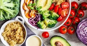 A bowl with assorted vegetables, quinoa, avocado, and cherry tomatoes, surrounded by more vegetables, quinoa, avocado, and a small bowl of dressing.