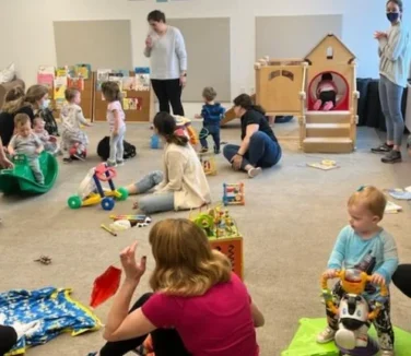 A group of young children and adults engage in play activities with toys in a classroom or daycare setting.