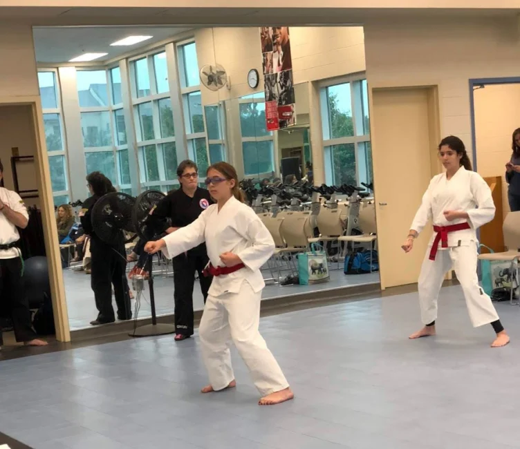 Two young people in white karate uniforms and red belts practice stances in a gym, while others watch from the background.