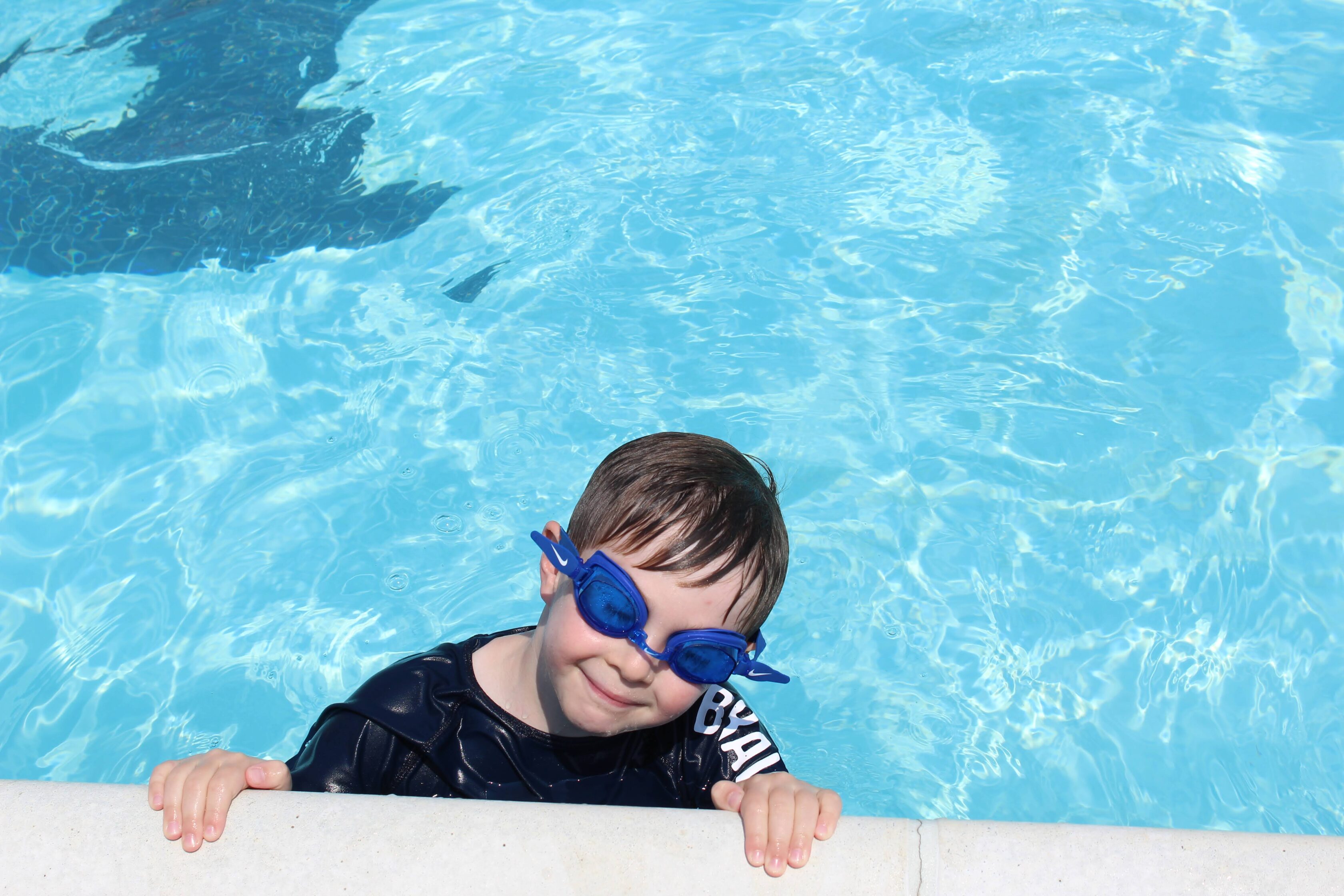 A young child wearing blue swim goggles holds onto the edge of a swimming pool, partially submerged in clear blue water.