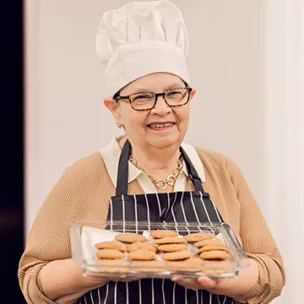 Older woman wearing a chef hat and apron holds a tray of freshly baked cookies, smiling at the camera in a kitchen setting.