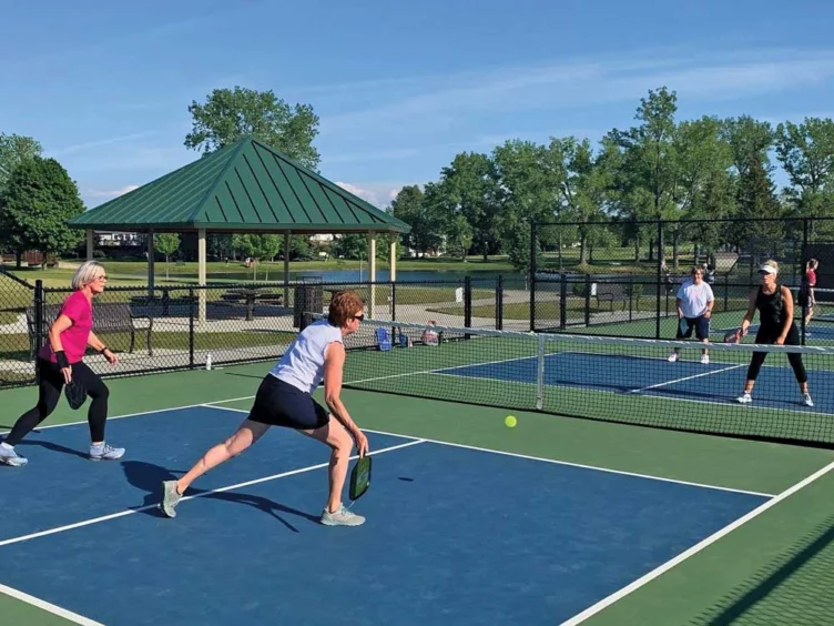 Four adults play a doubles pickleball match on an outdoor court, with a pavilion, trees, and a pond visible in the background.