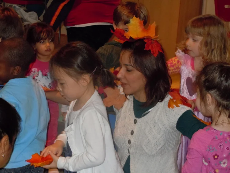 A woman with autumn leaves in her hair sits among several young children, some wearing costumes, in a classroom or daycare setting.