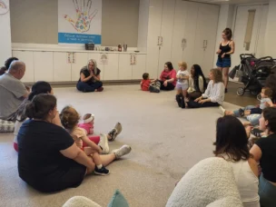 A group of adults and young children sit in a circle on the floor of a brightly lit room, listening to a woman who is speaking at the front.