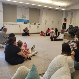 A group of adults and young children sit in a circle on the floor of a brightly lit room, listening to a woman who is speaking at the front.