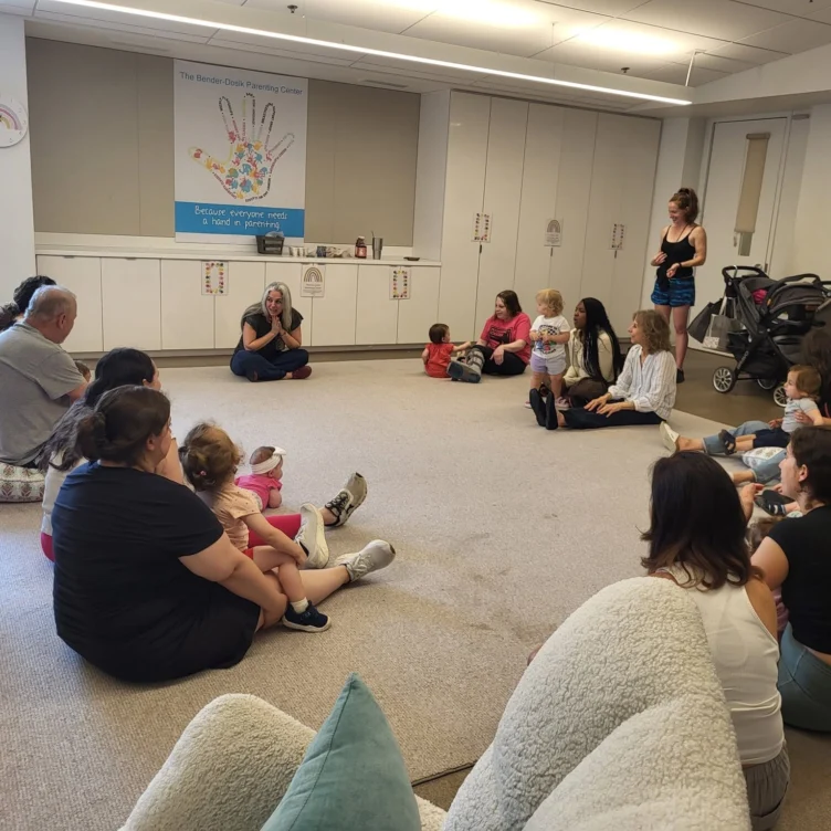 A group of adults and young children sit in a circle on the floor of a brightly lit room, listening to a woman who is speaking at the front.