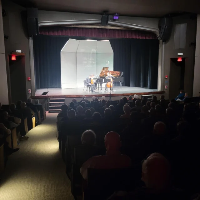 A chamber music trio performs on stage with a piano and string instruments in front of an audience in a dimly lit auditorium.