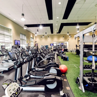 A spacious gym with treadmills, exercise bikes, weights, resistance balls, and turf flooring, well-lit with overhead lights and windows.