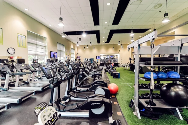 A spacious gym with treadmills, exercise bikes, weights, resistance balls, and turf flooring, well-lit with overhead lights and windows.