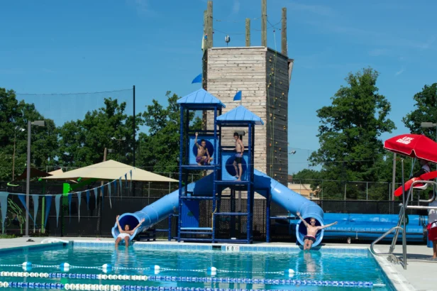 Four children use two blue slides that lead into an outdoor swimming pool, with a climbing tower structure and trees in the background.