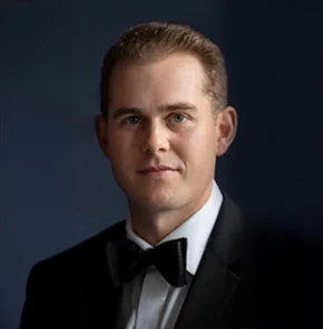 A man wearing a black tuxedo and bow tie poses for a formal portrait against a dark, blurred background.