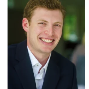 A young man with short, light brown hair is smiling while wearing a dark blazer and white shirt, seated indoors with a blurred background.