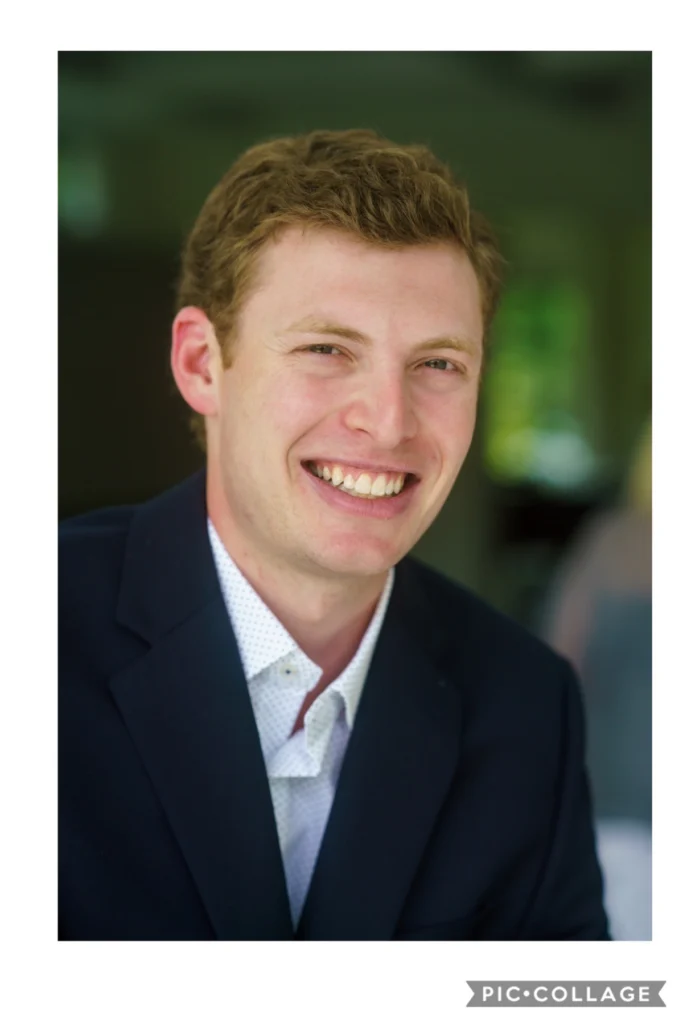 A young man with short, light brown hair is smiling while wearing a dark blazer and white shirt, seated indoors with a blurred background.