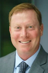 A man with short light brown hair wearing a gray suit, blue shirt, and patterned tie, smiling at the camera.