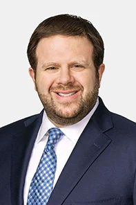 A man with short brown hair and a beard, wearing a navy blue suit, white shirt, and blue patterned tie, smiling in front of a light gray background.